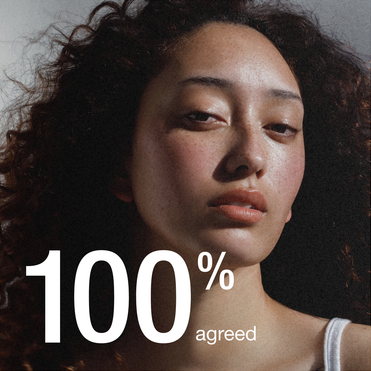 A close-up portrait of a woman with dark curly hair and a neutral expression, with large white text reading '100% agreed' overlaid across the lower portion.