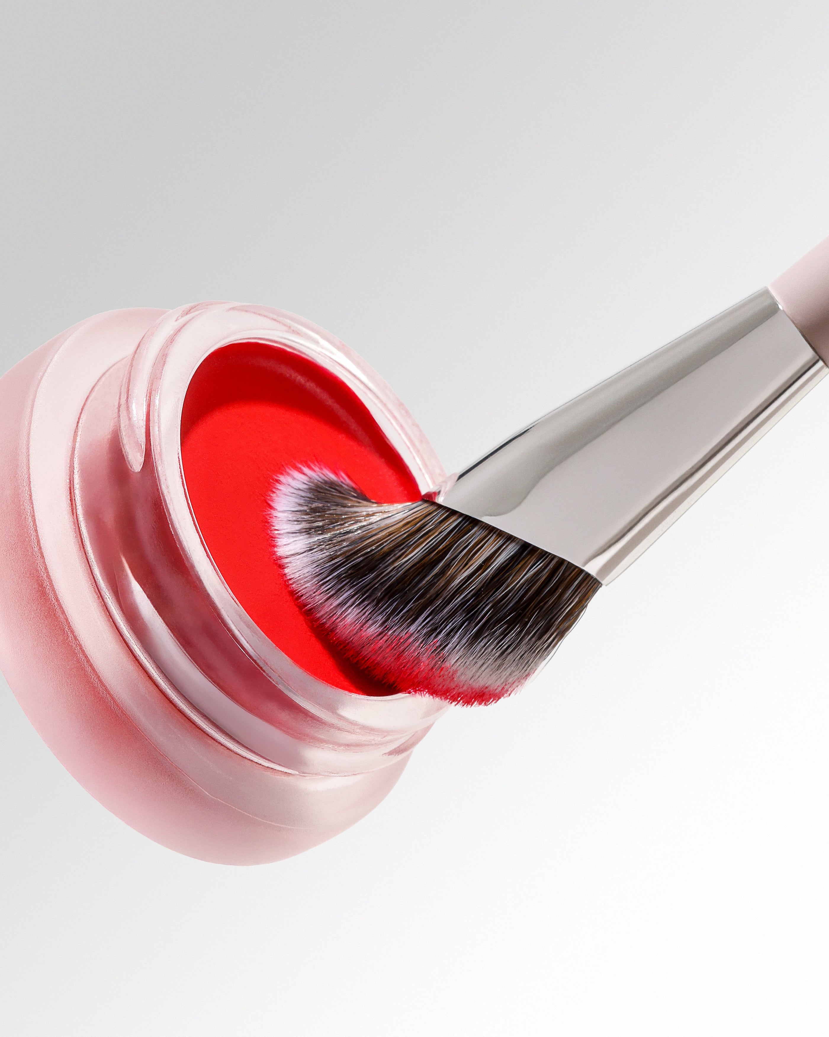 A close-up of a clear cosmetic jar filled with bright red cream, with a silver-handled brush dipped into the product against a light gray background.